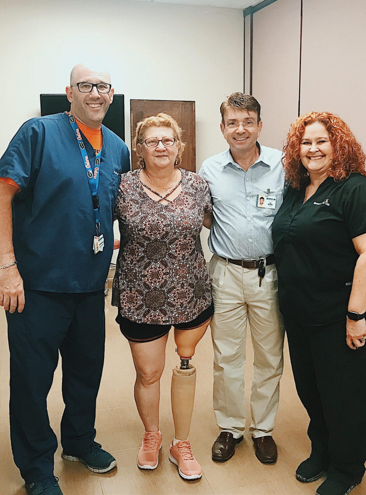 man in scrubs on far left, standing next to woman with prosthetic leg, another man on her right and a woman on his right. All are pictured smiling and posing for a picture in a clinic room.