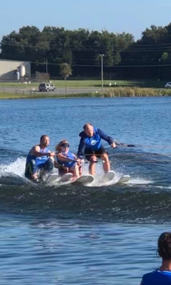 Without Limits adaptive water ski 2019 Woman smiling and waiving on an adaptive water ski, with one mane on either side holding her arms as they ski beside her. Amputee support group adaptive water skiing